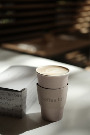 A tall, disposable coffee cup with a sleeve labeled 'coffee bar' is placed on a table with warm, soft lighting. In the background, blurred horizontal slats create a cozy and relaxed cafe atmosphere.
