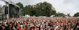 A large outdoor music festival with a packed crowd enjoying live performances. There is a stage on the left side with musicians performing, while the audience spans a vast area in front of the stage. Tall trees surround the event area, and there are white tents visible in the background, suggesting food or merchandise stalls. The atmosphere is lively with people raising their hands and cheering.