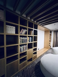A modern interior space features a large wooden bookshelf with several horizontal and vertical compartments. Some compartments contain a variety of books, while others are empty. The room has a patterned carpet on the floor and a round, white couch. The ceiling has wooden beams, adding to the room's contemporary design.