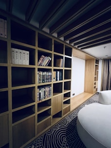 A modern interior space features a large wooden bookshelf with several horizontal and vertical compartments. Some compartments contain a variety of books, while others are empty. The room has a patterned carpet on the floor and a round, white couch. The ceiling has wooden beams, adding to the room's contemporary design.