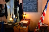 A vintage room showcasing historical artifacts, including an old-fashioned desk with a large open book, a lamp, and various containers on top. The wall features a chalkboard with writings, and an American flag stands beside a drum.
