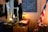 A vintage room showcasing historical artifacts, including an old-fashioned desk with a large open book, a lamp, and various containers on top. The wall features a chalkboard with writings, and an American flag stands beside a drum.