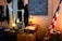 A vintage room showcasing historical artifacts, including an old-fashioned desk with a large open book, a lamp, and various containers on top. The wall features a chalkboard with writings, and an American flag stands beside a drum.