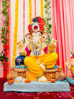 A colorful statue of the Hindu deity Ganesha is prominently displayed. The sculpture is adorned with vibrant jewelry and clothing, featuring a striking combination of yellow, pink, and red hues. Ganesha is seated with a pleasant expression, surrounded by decorative elements including flowers and green vines draping the background. The setting is festive and adorned with vertical garlands in the background.