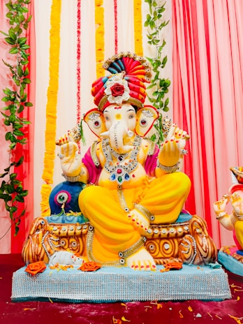 A colorful statue of the Hindu deity Ganesha is prominently displayed. The sculpture is adorned with vibrant jewelry and clothing, featuring a striking combination of yellow, pink, and red hues. Ganesha is seated with a pleasant expression, surrounded by decorative elements including flowers and green vines draping the background. The setting is festive and adorned with vertical garlands in the background.