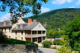 A large, historic house with a stone and brick exterior stands prominently on a lush green hillside. The architecture features gabled roofs, multiple chimneys, and a covered porch. A mountain covered with dense trees fills the background under a clear blue sky. A stone wall borders the property, and a curved road leads down the hill, surrounded by vibrant greenery.