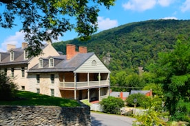 A large, historic house with a stone and brick exterior stands prominently on a lush green hillside. The architecture features gabled roofs, multiple chimneys, and a covered porch. A mountain covered with dense trees fills the background under a clear blue sky. A stone wall borders the property, and a curved road leads down the hill, surrounded by vibrant greenery.