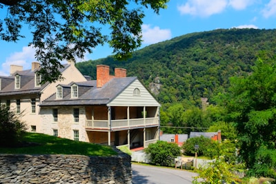 A large, historic house with a stone and brick exterior stands prominently on a lush green hillside. The architecture features gabled roofs, multiple chimneys, and a covered porch. A mountain covered with dense trees fills the background under a clear blue sky. A stone wall borders the property, and a curved road leads down the hill, surrounded by vibrant greenery.