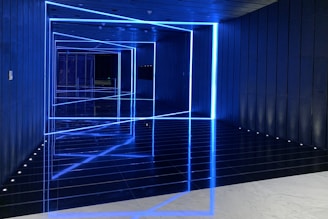The image features a futuristic hallway with blue neon lights outlining geometric shapes along the walls and ceiling, creating an optical illusion effect. The reflective floor enhances the symmetry and depth, adding to the modern and sleek appearance.