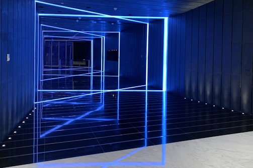 The image features a futuristic hallway with blue neon lights outlining geometric shapes along the walls and ceiling, creating an optical illusion effect. The reflective floor enhances the symmetry and depth, adding to the modern and sleek appearance.
