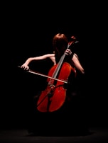 A person playing a cello in a dimly lit scene, with the focus on the warm, reddish-brown color of the cello against a dark background. The musician's features are obscured by shadows, creating a dramatic and mysterious atmosphere.