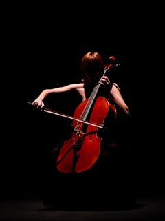 A person playing a cello in a dimly lit scene, with the focus on the warm, reddish-brown color of the cello against a dark background. The musician's features are obscured by shadows, creating a dramatic and mysterious atmosphere.