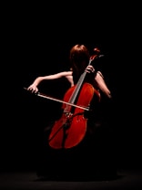 A person playing a cello in a dimly lit scene, with the focus on the warm, reddish-brown color of the cello against a dark background. The musician's features are obscured by shadows, creating a dramatic and mysterious atmosphere.