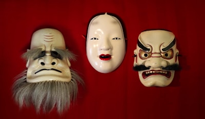 Three traditional Japanese masks are displayed side by side on a red background. The mask on the left features exaggerated eyebrows and a long, gray beard, conveying an expression of wisdom or grumpiness. The center mask depicts a serene and elegant female face with pale skin and red lips, evoking a sense of calm beauty. The mask on the right shows a fierce expression with furrowed brows, red accents, and a black mustache, suggesting intensity or anger.