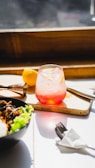 A refreshing pink drink is served in a frosted glass with ice and a slice of citrus on a wooden tray near a window that lets in bright sunlight. Beside it, a yellow lemon, a copper jigger, and a bowl of food with fresh green lettuce and cooked meat are visible. A napkin and fork rest on the white surface.