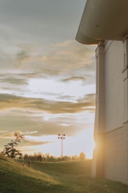 A serene landscape featuring a partially cloudy sky with soft golden light, a large building rising from the right side of the frame. A lamppost and sparse trees are scattered over a grassy hill in the foreground, bathed in the warm glow of the setting sun.