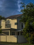 A modern, two-story house with clean, minimalist architecture is partially obscured by lush green trees. The building features large windows and white exterior walls. The sky is clear with a few wispy clouds, suggesting calm weather.
