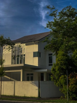 A modern, two-story house with clean, minimalist architecture is partially obscured by lush green trees. The building features large windows and white exterior walls. The sky is clear with a few wispy clouds, suggesting calm weather.
