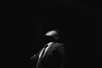 A man in a suit stands in a dark setting with a spotlight illuminating his face and upper body. His head is tilted slightly upwards with a contemplative or serene expression.