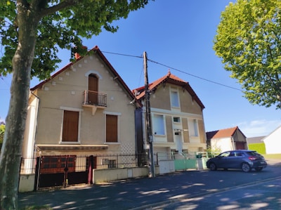 Two residential houses with red-tiled roofs are seen under a clear blue sky. The buildings have a classic European architectural style with shutters on the windows. A car is parked on the paved street in front of the houses. Trees with lush green foliage frame the scene, casting shadows on the ground.