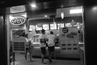 A black and white image of a fast-food restaurant with a glass entrance door. There are three people standing inside at the counter, seemingly placing an order. An 'Open' sign is lit above the entrance, and promotional posters are visible on the glass, advertising specials. Inside, the counter displays menus and options typically found in a fast-food establishment.