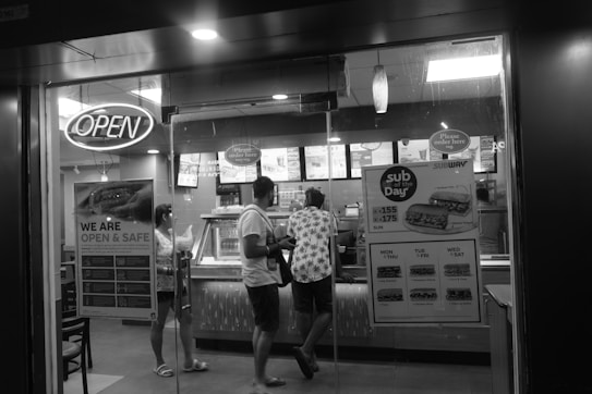 A black and white image of a fast-food restaurant with a glass entrance door. There are three people standing inside at the counter, seemingly placing an order. An 'Open' sign is lit above the entrance, and promotional posters are visible on the glass, advertising specials. Inside, the counter displays menus and options typically found in a fast-food establishment.
