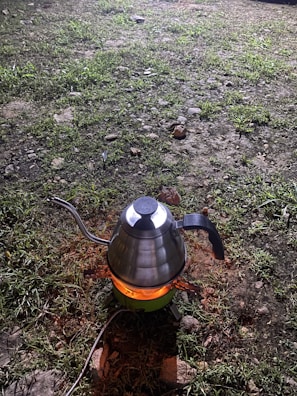 A metallic kettle with a curved spout and black handle is placed on a small portable stove. The stove is emitting an orange glow, indicating it is lit. The surrounding area is covered in grass and small rocks, suggesting an outdoor setting, likely at night due to the surrounding darkness and artificial light source highlighting the kettle.