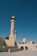 A mosque featuring tall minarets and domes constructed from beige stone. The architectural style reflects traditional Islamic designs, with archways and ornamental details. The surrounding area is open with a clear blue sky overhead.