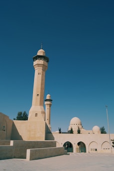 A mosque featuring tall minarets and domes constructed from beige stone. The architectural style reflects traditional Islamic designs, with archways and ornamental details. The surrounding area is open with a clear blue sky overhead.