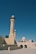 A mosque featuring tall minarets and domes constructed from beige stone. The architectural style reflects traditional Islamic designs, with archways and ornamental details. The surrounding area is open with a clear blue sky overhead.