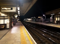 A dimly lit train station platform with empty tracks extending into the distance. The platform features a clock, benches, and signs, including one indicating 'Way out'. Overhead lights illuminate the area, highlighting the safety yellow lines along the edges of the platform.