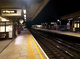 A dimly lit train station platform with empty tracks extending into the distance. The platform features a clock, benches, and signs, including one indicating 'Way out'. Overhead lights illuminate the area, highlighting the safety yellow lines along the edges of the platform.