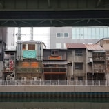 A row of older, slightly worn buildings with air conditioning units visible on their facades. One building has a sign 'La Oliva' and another has a teal sheet hanging. There is a modern building in the background. The scene is viewed from across a water canal, under a bridge or overhead structure.