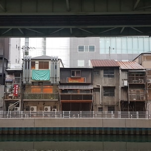 A row of older, slightly worn buildings with air conditioning units visible on their facades. One building has a sign 'La Oliva' and another has a teal sheet hanging. There is a modern building in the background. The scene is viewed from across a water canal, under a bridge or overhead structure.