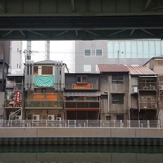 A row of older, slightly worn buildings with air conditioning units visible on their facades. One building has a sign 'La Oliva' and another has a teal sheet hanging. There is a modern building in the background. The scene is viewed from across a water canal, under a bridge or overhead structure.