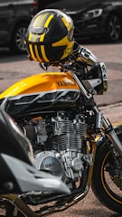 A close-up view of a yellow and black motorcycle parked on a street. The focus is on the engine and body of the Yamaha motorcycle, with a coordinated helmet resting on the handlebars. The background shows blurred vehicles, emphasizing the motorcycle.