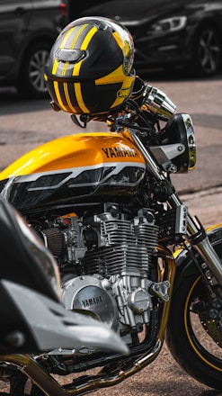 A close-up view of a yellow and black motorcycle parked on a street. The focus is on the engine and body of the Yamaha motorcycle, with a coordinated helmet resting on the handlebars. The background shows blurred vehicles, emphasizing the motorcycle.
