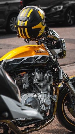 A close-up view of a yellow and black motorcycle parked on a street. The focus is on the engine and body of the Yamaha motorcycle, with a coordinated helmet resting on the handlebars. The background shows blurred vehicles, emphasizing the motorcycle.