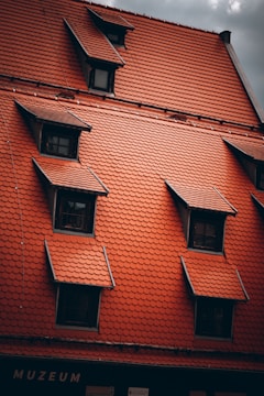 A red-tiled roof with multiple windows and dormers, featuring a traditional architectural style. The tiles are arranged in an orderly fashion, complementing the structure underneath.