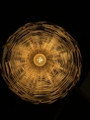 A circular woven lampshade with a warm light bulb in the center, casting intricate shadows and patterns on the inside surfaces.