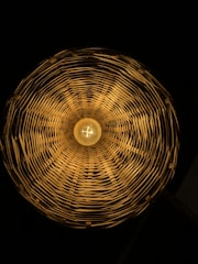 A circular woven lampshade with a warm light bulb in the center, casting intricate shadows and patterns on the inside surfaces.