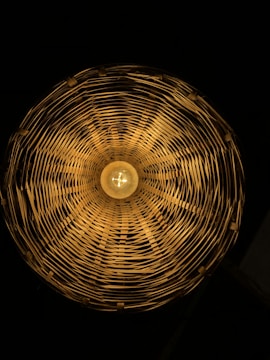 A circular woven lampshade with a warm light bulb in the center, casting intricate shadows and patterns on the inside surfaces.
