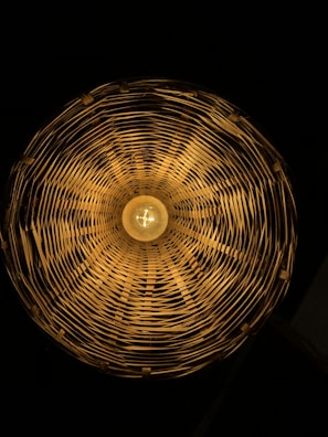 A circular woven lampshade with a warm light bulb in the center, casting intricate shadows and patterns on the inside surfaces.
