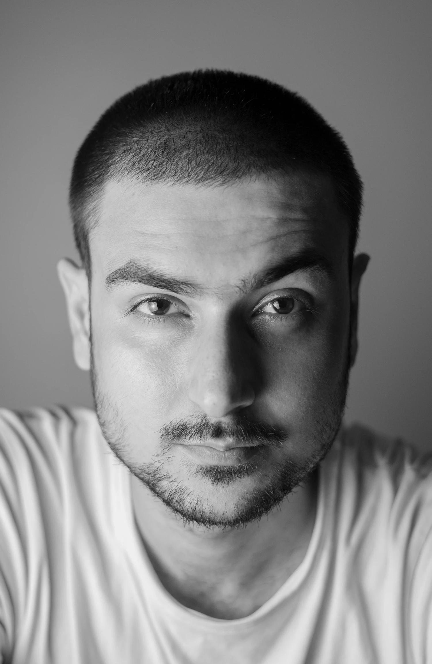 A man in a plain white t-shirt against a light grey wall, photographed head-on