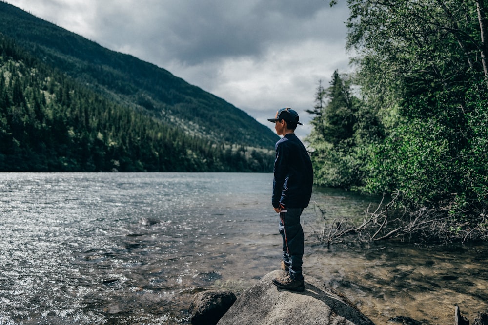 a man standing on a rock by a river with trees and mountains in the background