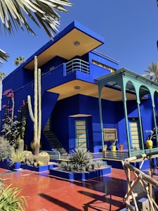A vibrant blue building with yellow accents stands amidst a lush garden. Tall cacti and various plants decorate the entrance, while a green and blue pergola provides shade. The architecture features clean lines and geometric shapes, offering a modern aesthetic enhanced by the striking color contrast against the clear blue sky.