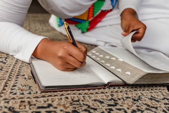 A person is writing in a notebook while another book is open nearby, placed on a patterned carpet. The individual is dressed in white, with colorful beaded decorations visible on their clothing. One hand is holding a pen, and the other is turning a page in the book.