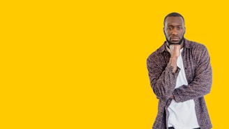 A man stands confidently against a solid yellow background, wearing a patterned dark shirt over a white t-shirt. He has his chin resting on one hand, exuding a thoughtful yet assured expression.