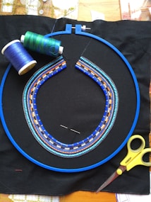 A beaded necklace design in progress is displayed within a blue embroidery hoop on black fabric. The design features intricate patterns in shades of blue, orange, and white beads. Nearby are spools of green and blue thread, a needle, and a pair of yellow scissors, suggesting an ongoing beadwork or embroidery project.