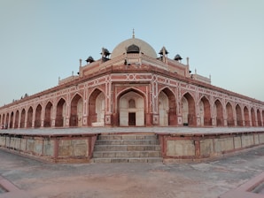 A large, symmetrical red sandstone building with detailed Mughal architecture, featuring a dome and arched entrances. The structure has intricate white inlay work on its facade and is set on a raised platform with stone steps leading up to it.